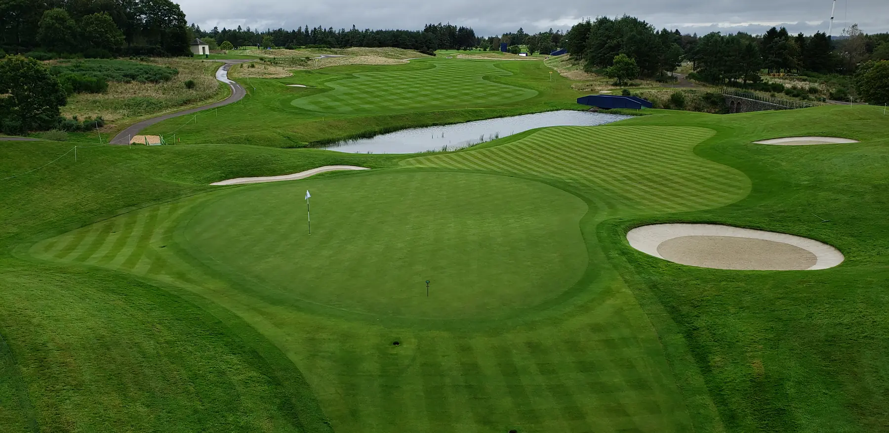 Aerial view of a complete golf hole: a flagged green in the foreground, bunkers, a pond and a striped fairway running back to the treeline, with a TOMi instrument standing on the green