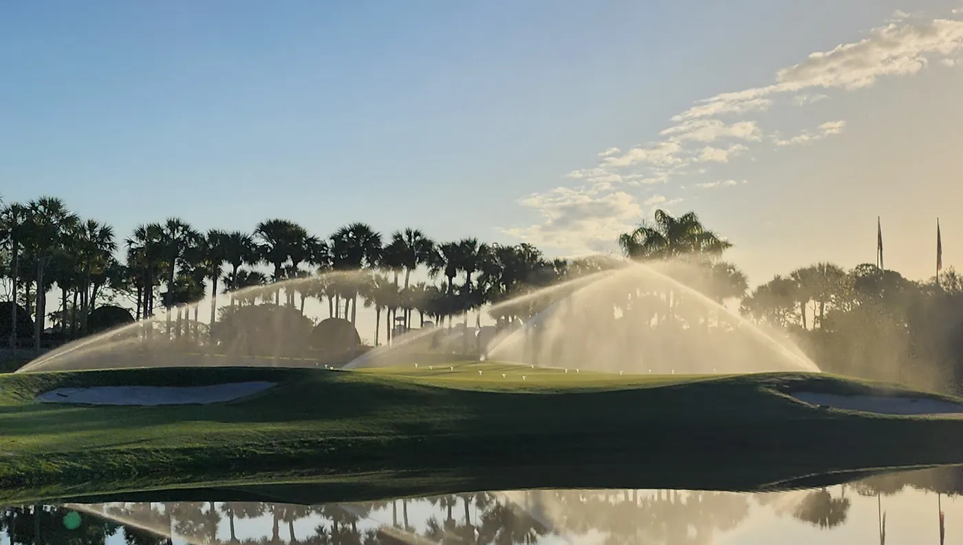 Sunrise over a palm-lined golf course, irrigation heads throwing arcs of water across the green and the whole scene mirrored in the pond in front of it