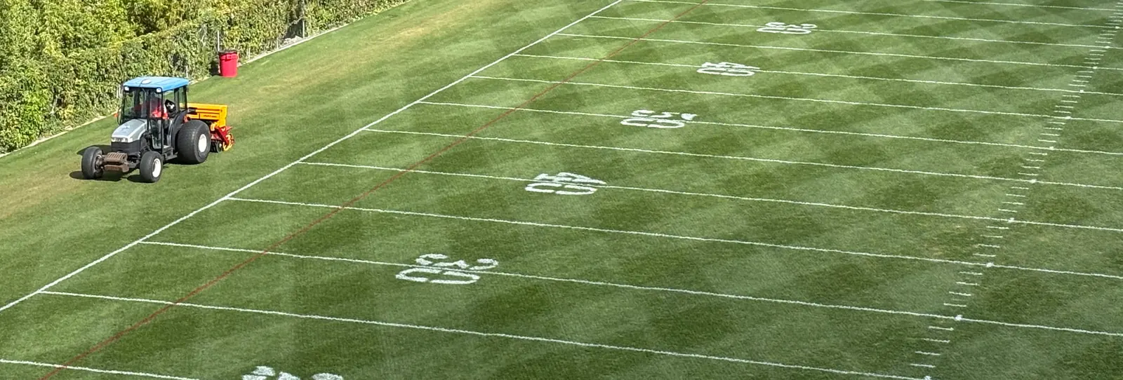 A compact tractor towing a seeder along the sideline of a marked American football field in bright sun