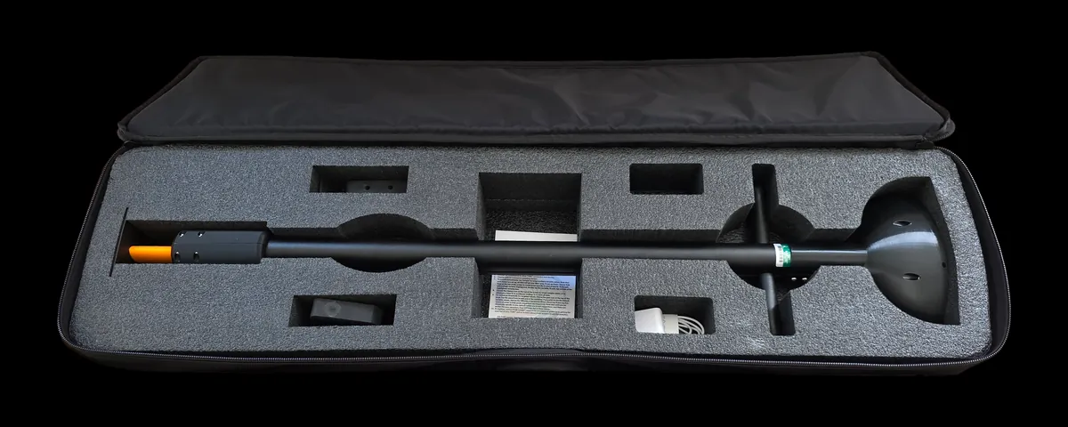 The TOMi instrument lying in its fitted foam carry case: the shaft with its orange-capped sensor tip, crossbar handle and base plate seated in cut-outs, spare accessories and a printed guide in the smaller recesses, and the padded lid folded open behind