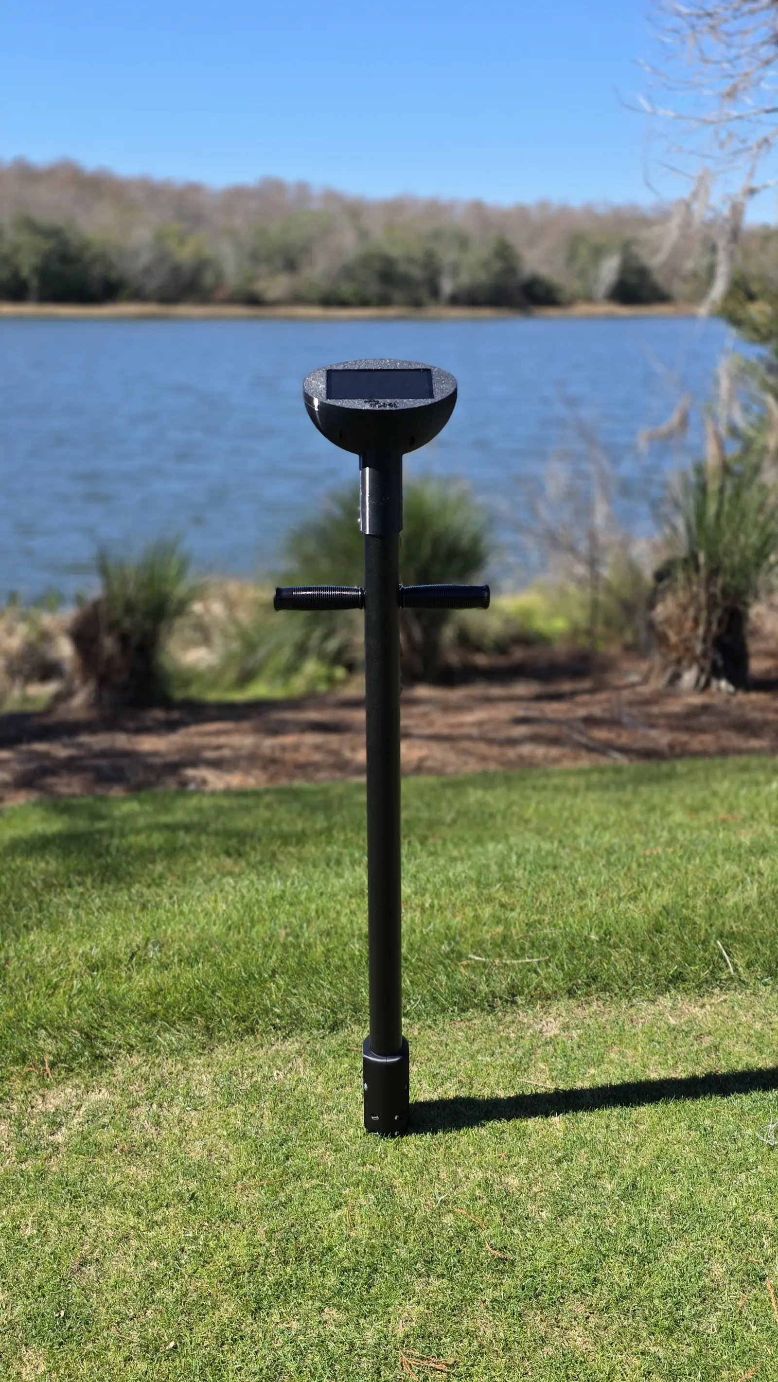 A TOMi instrument standing upright in mown turf, its solar-topped head and handles clear against a pond and treeline behind