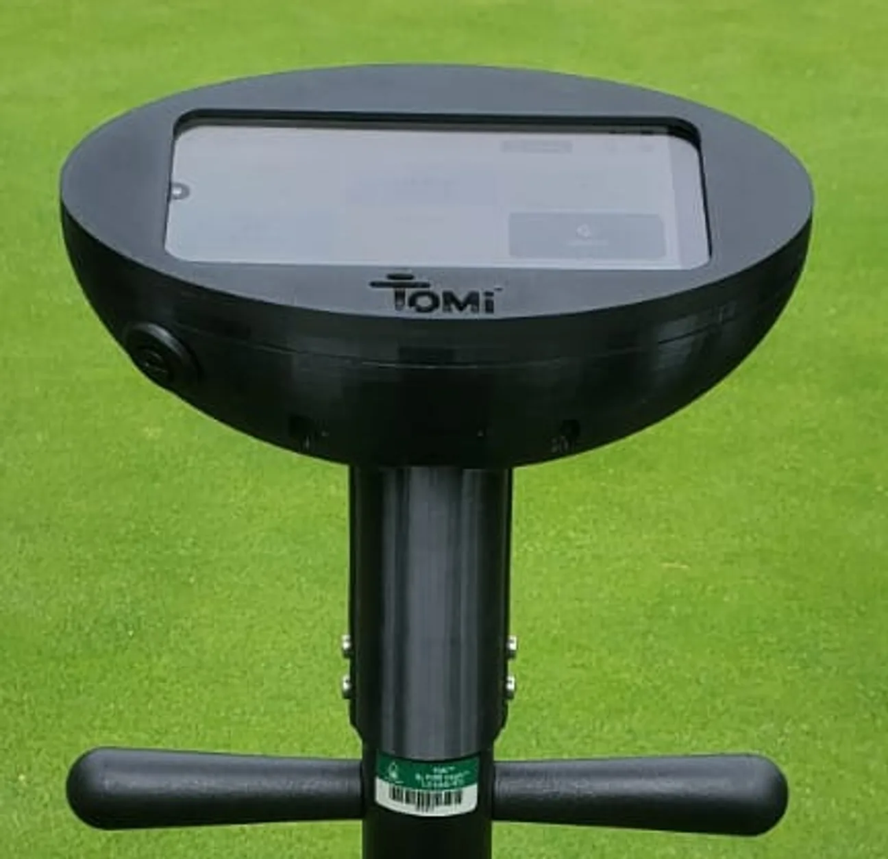 A TOMi standing in closely mown turf, photographed from the operator&rsquo;s eye line: the black oval head with its landscape touchscreen set into the top face, the shaft below it and the handle bars across it